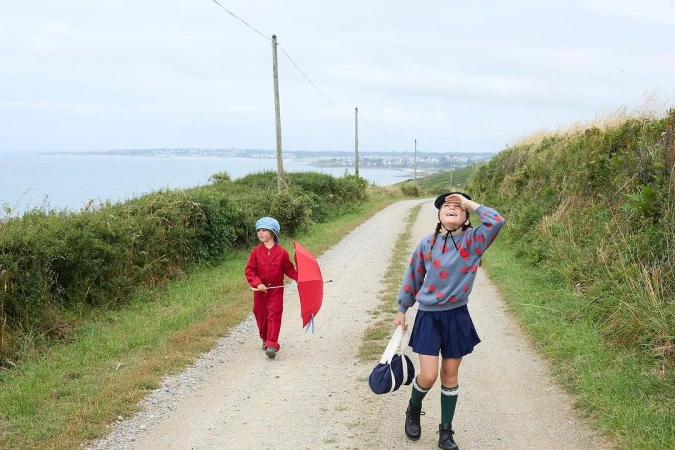 Two children, one with an umbrella, walk down a path on a coastline with the ocean and coastal town in the background on an overcast day.