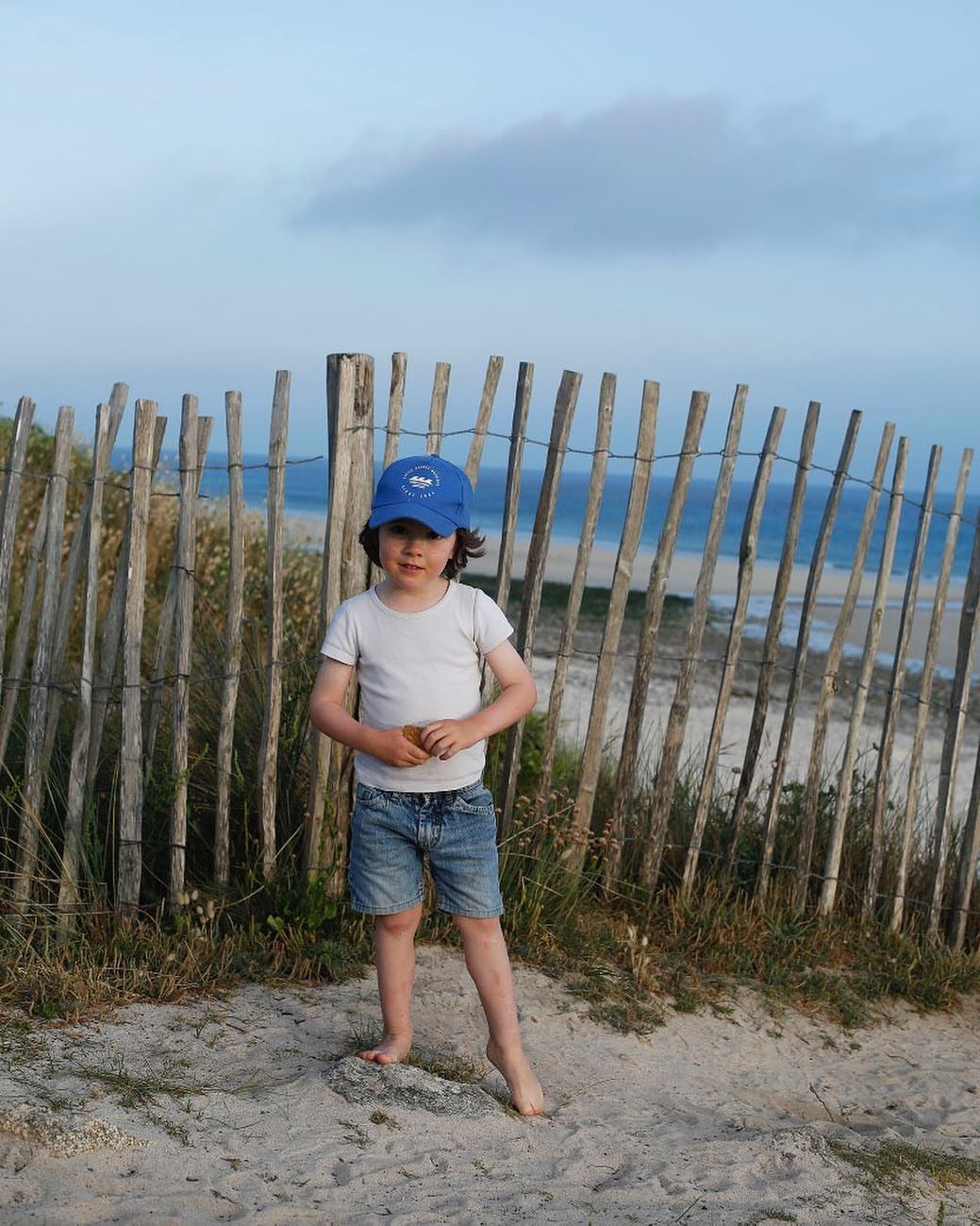 Young child standing barefoot on a sandy beach near a wooden fence with a blue cap, white t-shirt, and denim shorts, with ocean view.