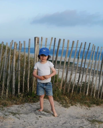 Child wearing a blue cap, white t-shirt, and denim shorts standing on sandy ground in front of a wooden fence and ocean.