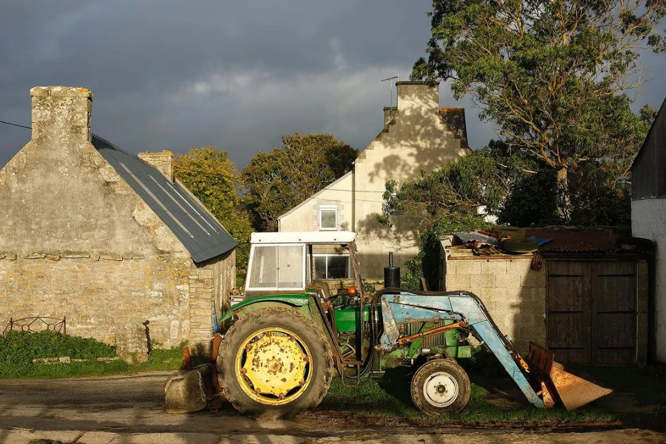 Weathered green and blue tractor with yellow wheels parked in a rural setting next to stone buildings and a cloudy sky.