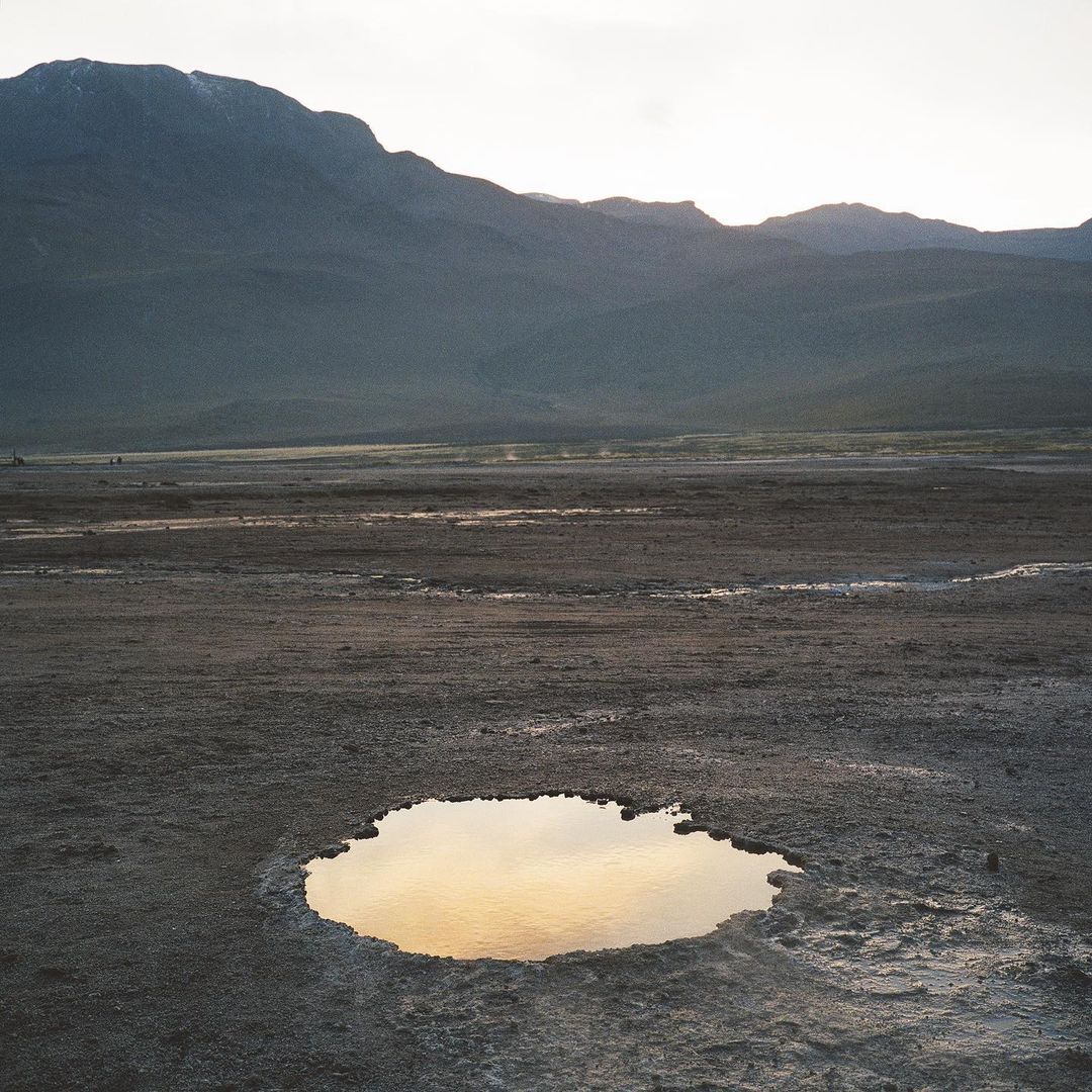Desert landscape with mountains and a small pool of water reflecting the sky at dusk, creating a serene and minimalist natural scene.