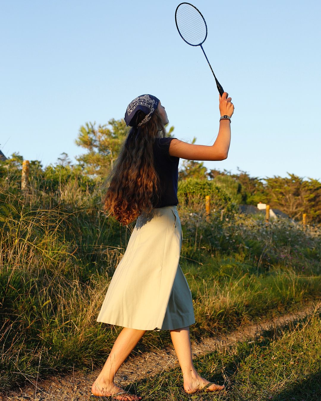 Young woman plays badminton outdoors in natural light. She wears a bandana, t-shirt, and skirt, standing on a grassy path surrounded by greenery.