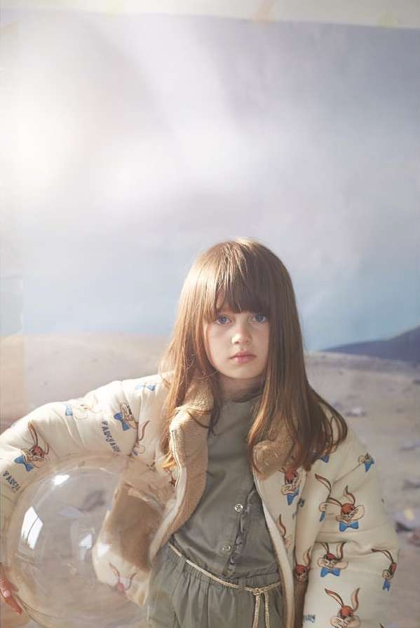 Young girl with long brown hair wearing a printed jacket and holding a clear bubble in an indoor setting with a painted background.