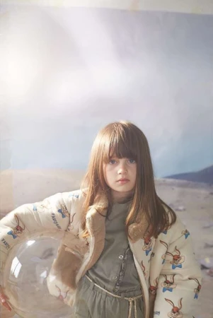 Young girl with long brown hair wearing a printed jacket and holding a clear bubble in an indoor setting with a painted background.