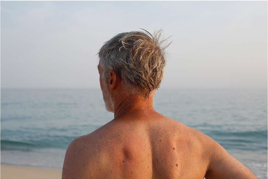 Rear view of an older man with grey hair looking out toward the ocean from a sandy beach at sunset.