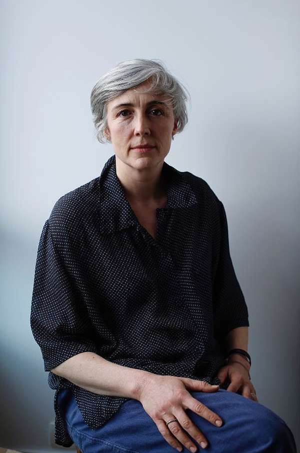 Portrait of a silver-haired woman in a black and white polka dot blouse and blue jeans sitting against a white wall, looking at the camera.