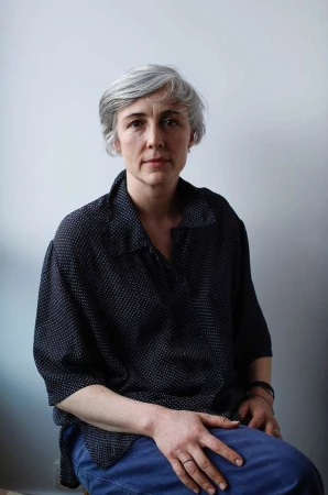 Portrait of a silver-haired woman in a black and white polka dot blouse and blue jeans sitting against a white wall, looking at the camera.