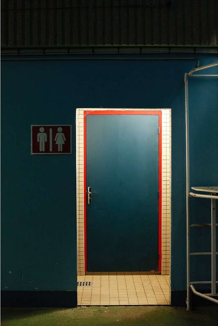 Illuminated doorway in a blue-walled building, indicated as a public restroom with a sign featuring male and female figures.