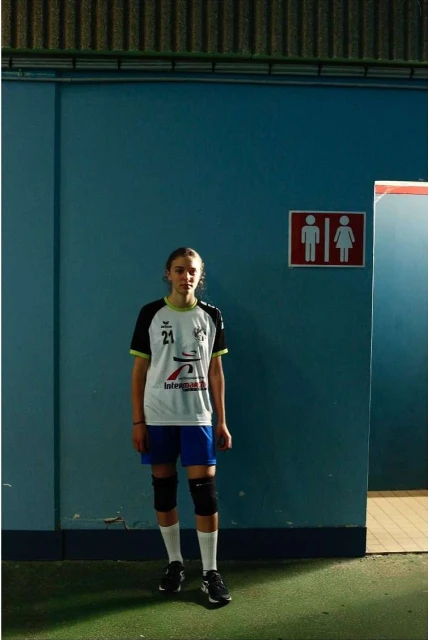 Teenage girl athlete in sportswear stands in front of a blue indoor wall near a restroom sign, in a medium studio shot.