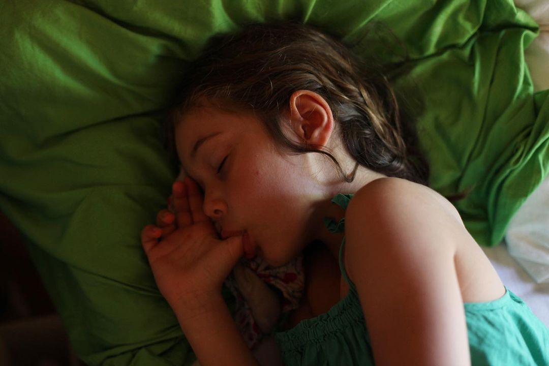 Young girl sleeping on a green pillow and white bedding, resting her face on her hand, wearing a green top.