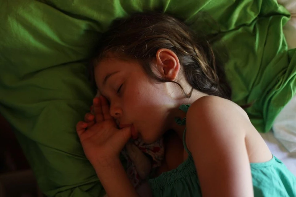 Young girl sleeping on a green pillow and white bedding, resting her face on her hand, wearing a green top.