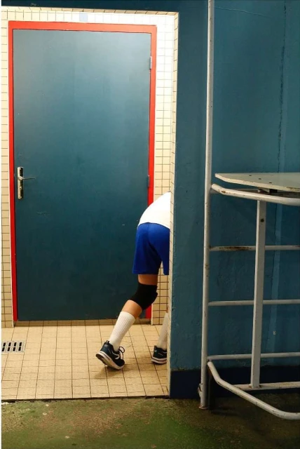 Person emerging from a blue door in a tiled room, wearing blue shorts, white socks, and black sneakers, with a metallic table nearby.