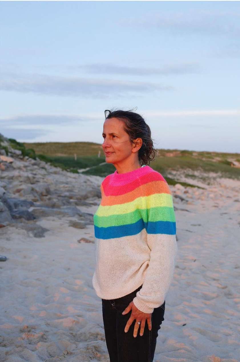 Adult woman standing on a beach, wearing a rainbow sweater, with a beach and grassy dunes in the soft evening light.