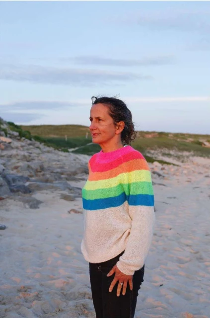 Adult woman standing on a beach, wearing a rainbow sweater, with a beach and grassy dunes in the soft evening light.