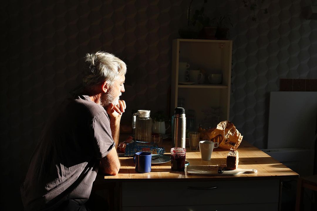 Elderly man with gray hair sits at a sunlit table with food, jam, a blue mug, and a silver thermos in an indoor setting.