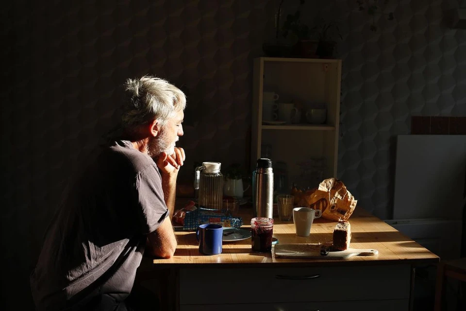 Elderly man with gray hair sits at a sunlit table with food, jam, a blue mug, and a silver thermos in an indoor setting.