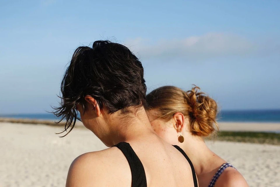 Two adult females stand on a beach with light sand and calm, blue ocean waters under a clear sky during daylight hours.