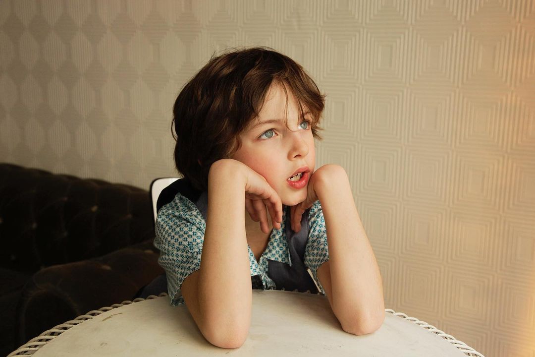 Child with brown hair poses at table indoors, looking upwards with patterned wallpaper and brown sofa in the background.