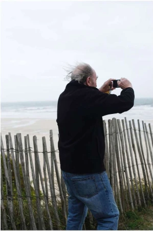 Elderly man in a dark jacket and blue jeans standing behind a wooden fence, taking a photo with his smartphone near the ocean.