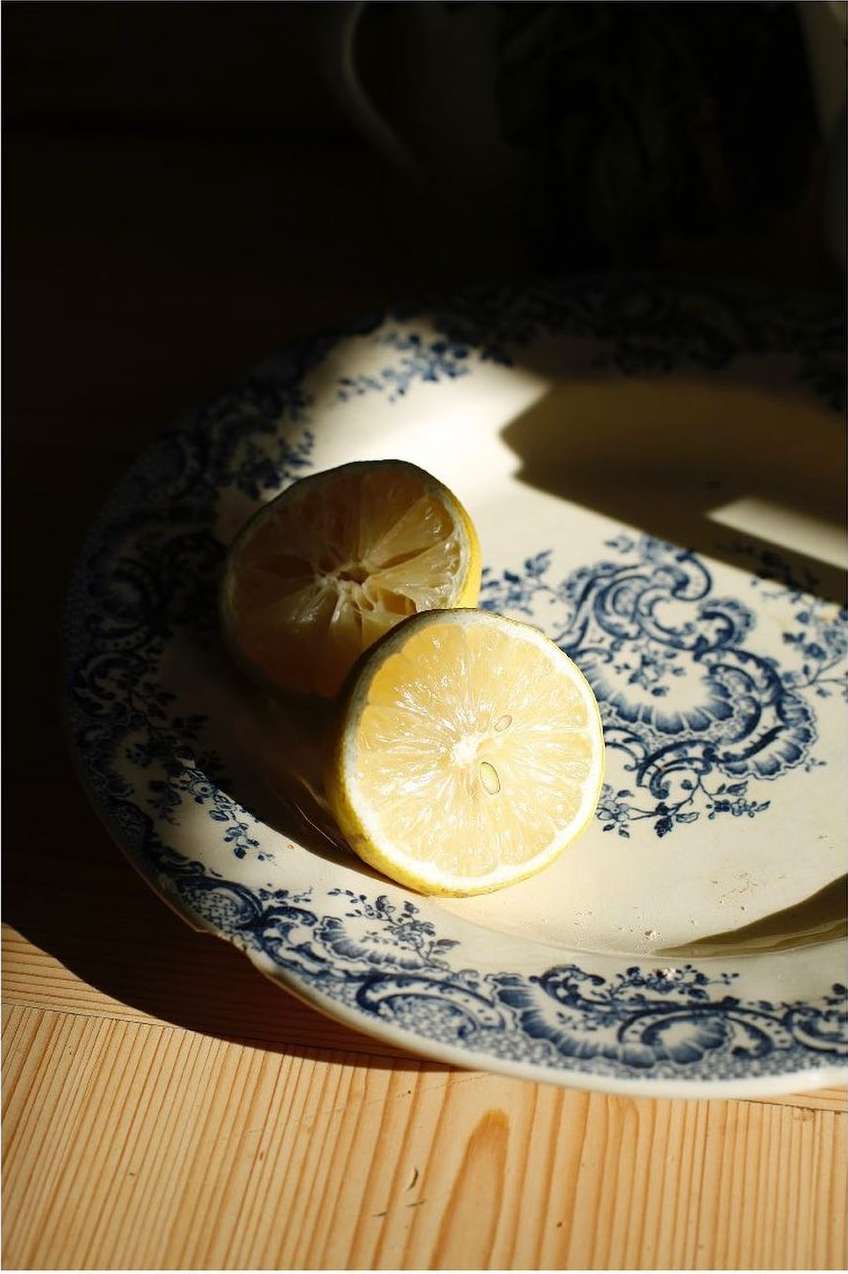 Still life photograph of two lemon halves on an ornamental plate on a wooden surface, under dramatic lighting.