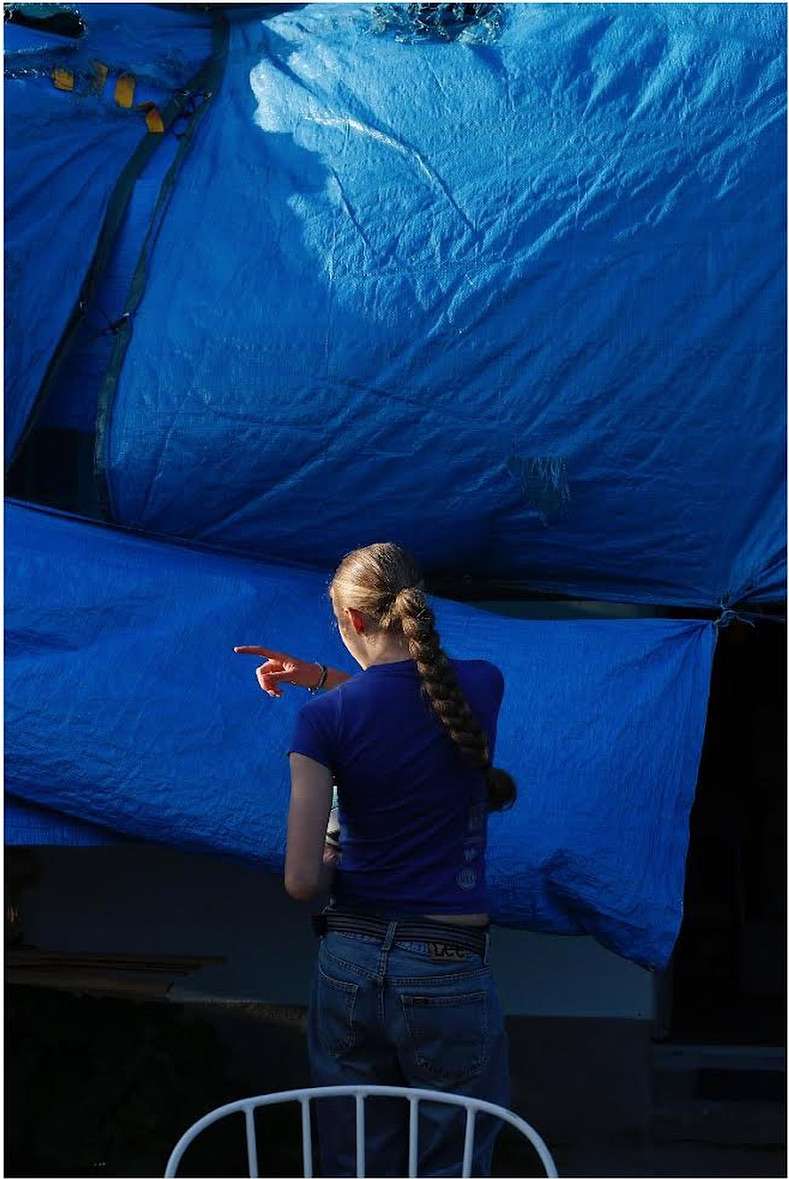 A young woman with a braid standing in a blue shirt and jeans points toward a large, blue tarp overhead in an outdoor setting.