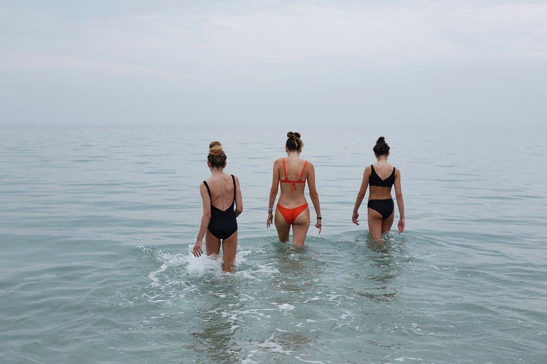 Three women in swimsuits walk into the ocean on a cloudy day. Back view of women in bathing suits wading into water.