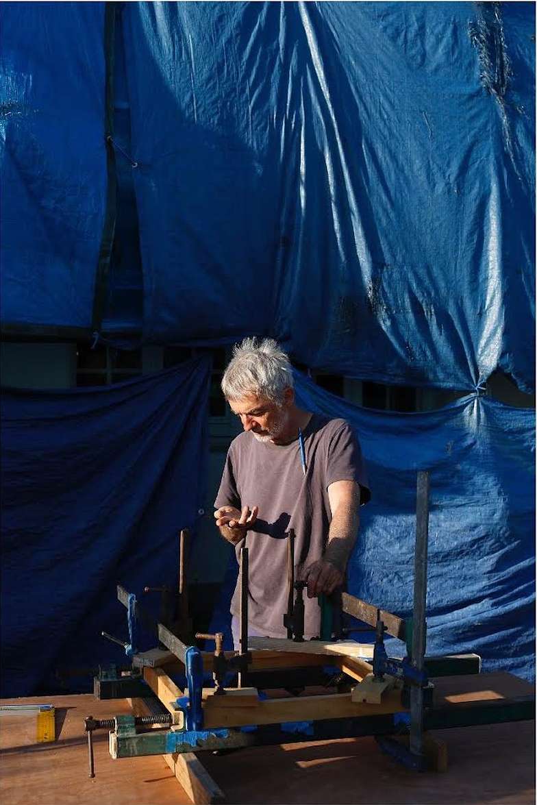 Adult man working on a wood construction project outdoors on a workbench, using clamps under a blue tarp during the daytime.