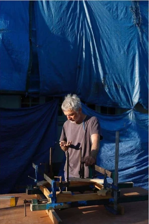 Adult man working on a wood construction project outdoors on a workbench, using clamps under a blue tarp during the daytime.