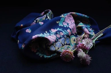 A deep blue floral silk scarf draped next to a small bunch of pink flowers against a black background