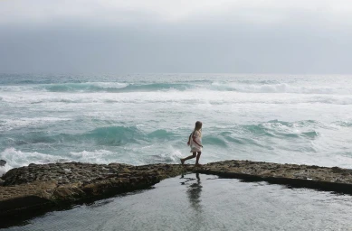 A young girl walking along a rocky edge by turbulent ocean waves under an overcast sky, captured by photographer Camille Malissen.