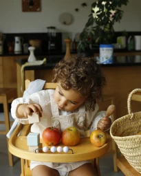 A toddler with curly hair seated in a wooden high chair playing with wooden blocks and apples.