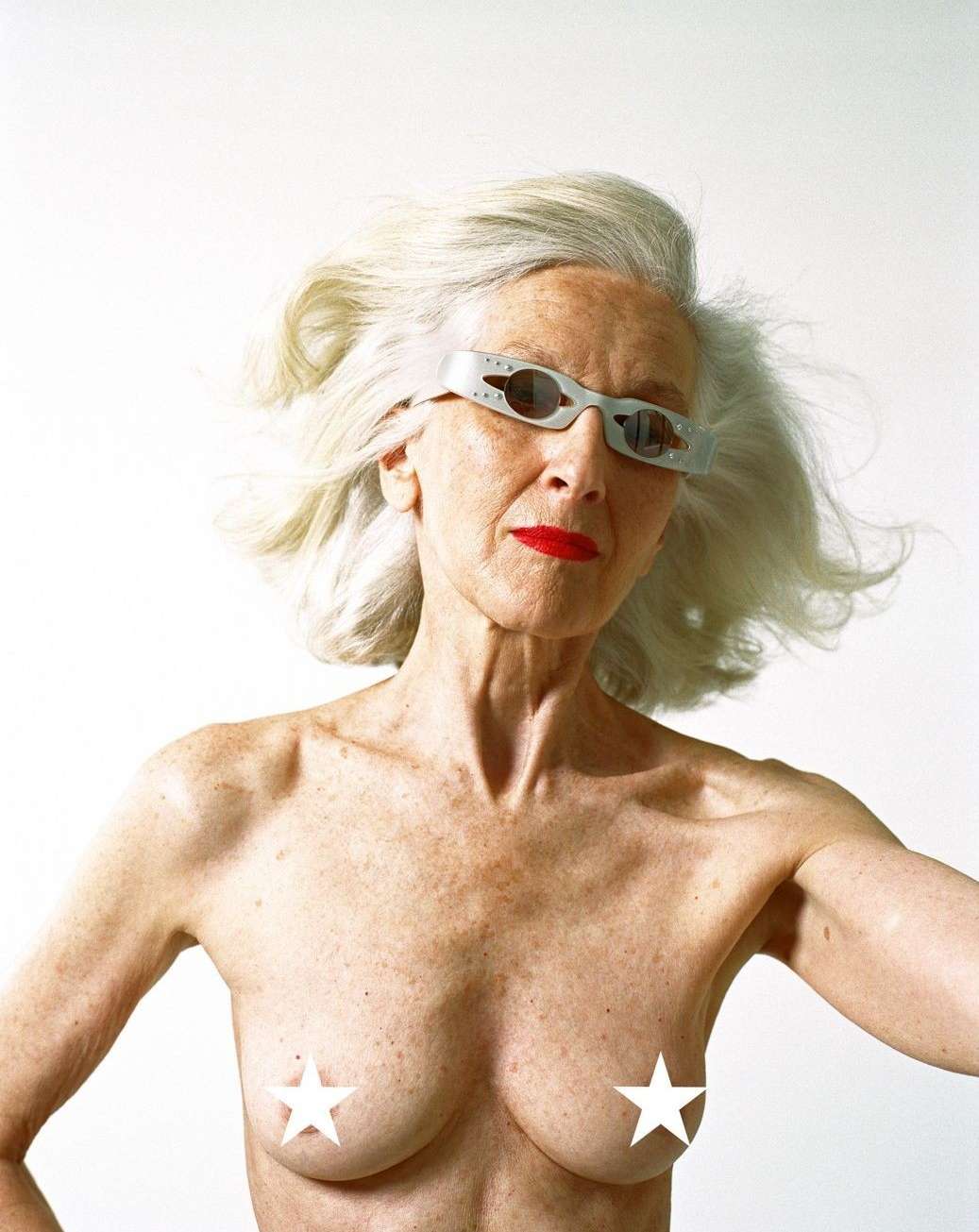 Older woman with silver hair, bright red lipstick, and unique glasses poses in front of a white backdrop in a studio setting.