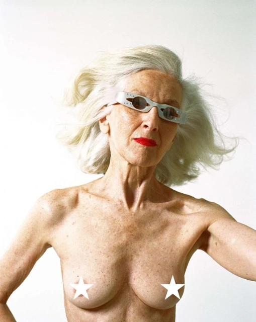 Older woman with silver hair, bright red lipstick, and unique glasses poses in front of a white backdrop in a studio setting.