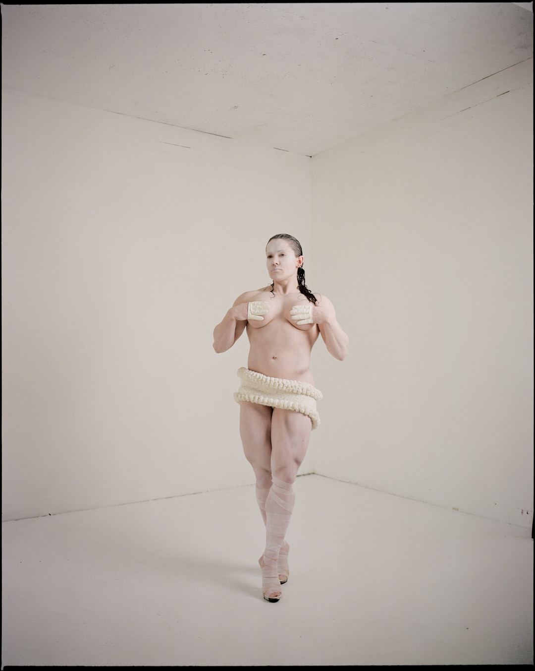 Athletic adult female covered in white paint poses in empty white corner room wearing knitted wrap and platform shoes, looking at the camera.