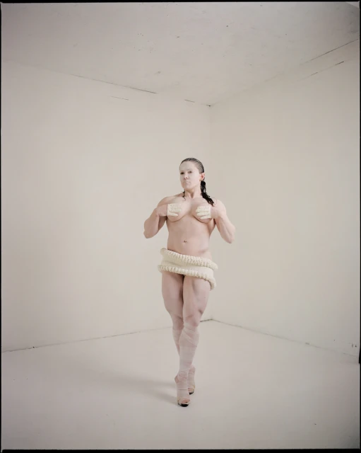 Athletic adult female covered in white paint poses in empty white corner room wearing knitted wrap and platform shoes, looking at the camera.