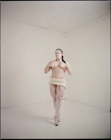 Athletic adult female covered in white paint poses in empty white corner room wearing knitted wrap and platform shoes, looking at the camera.