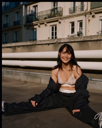 Smiling woman in lace bralette and loose dark trousers sitting outdoors against a Parisian building exterior