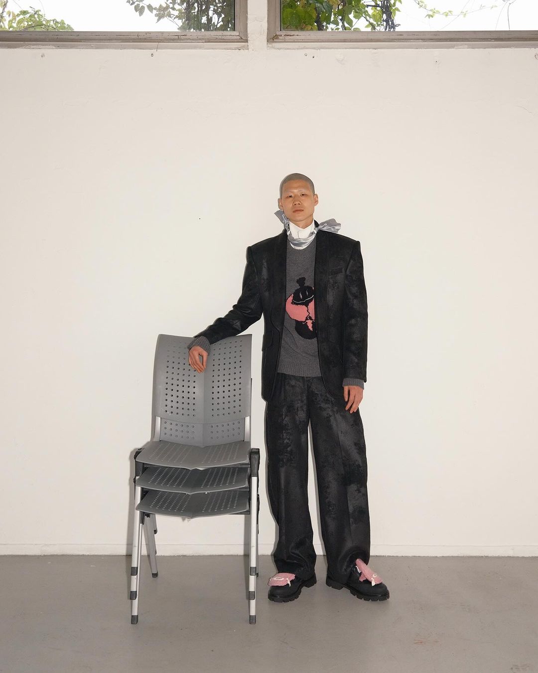 A person with a shaved head poses indoors in a dark patterned suit, grey sweater, and platform shoes, standing next to stacked gray chairs.