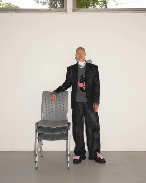 A person with a shaved head poses indoors in a dark patterned suit, grey sweater, and platform shoes, standing next to stacked gray chairs.
