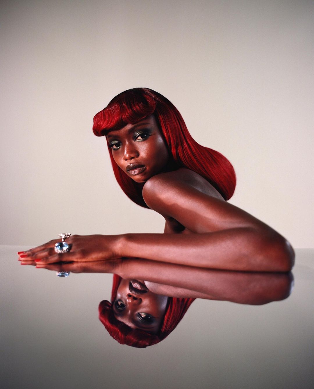 Elegant portrait of a Black woman with red hair and a large blue ring, reflected on a glossy surface against a pale background.