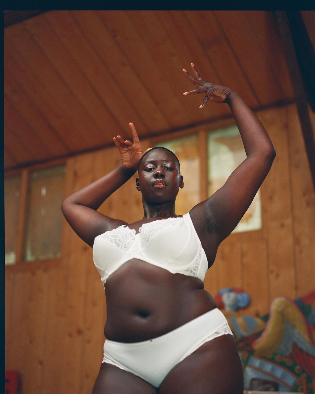 A woman poses in a white bra and underwear in front of a paneled wall with her arms raised in an indoor setting.