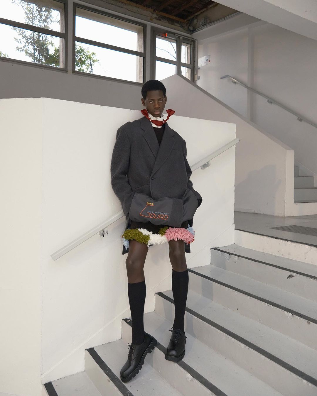 Fashionable Black male model posing on stairs in an oversized grey blazer, black knee socks, and leather shoes, showcasing avant-garde style.
