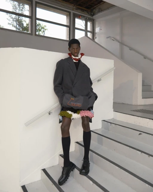 Fashionable Black male model posing on stairs in an oversized grey blazer, black knee socks, and leather shoes, showcasing avant-garde style.