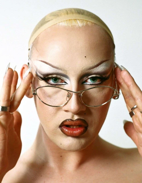 Close-up portrait of a person with light skin, heavy makeup, glasses, and long nails, against a white background.