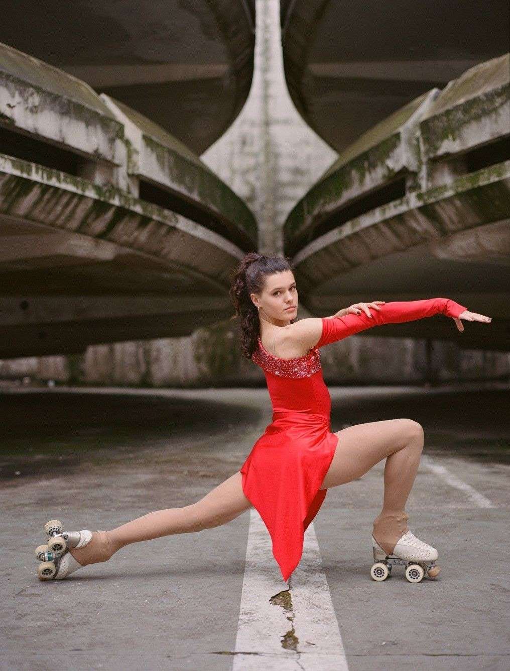 A woman in a red dress on roller skates in a performing pose against a grey, outdoor, architectural background.