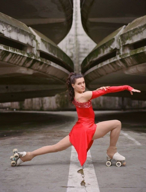 A woman in a red dress on roller skates in a performing pose against a grey, outdoor, architectural background.