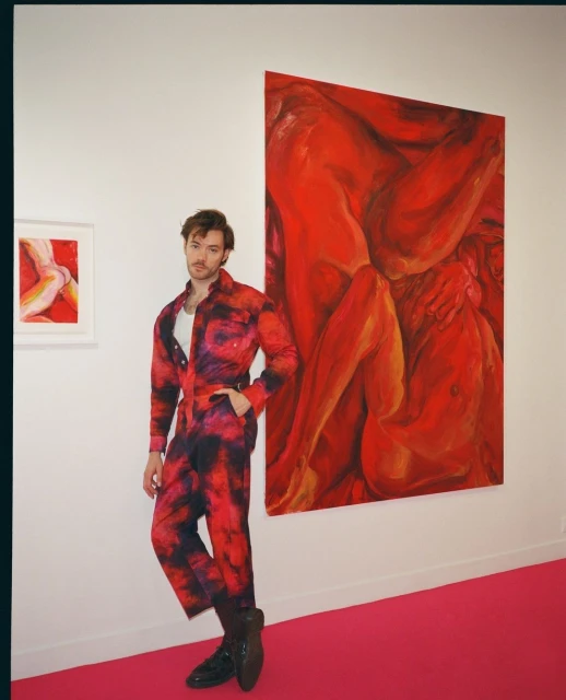 Man in patterned jumpsuit stands in gallery next to red abstract paintings and pink carpet, showcasing contemporary art and fashion.