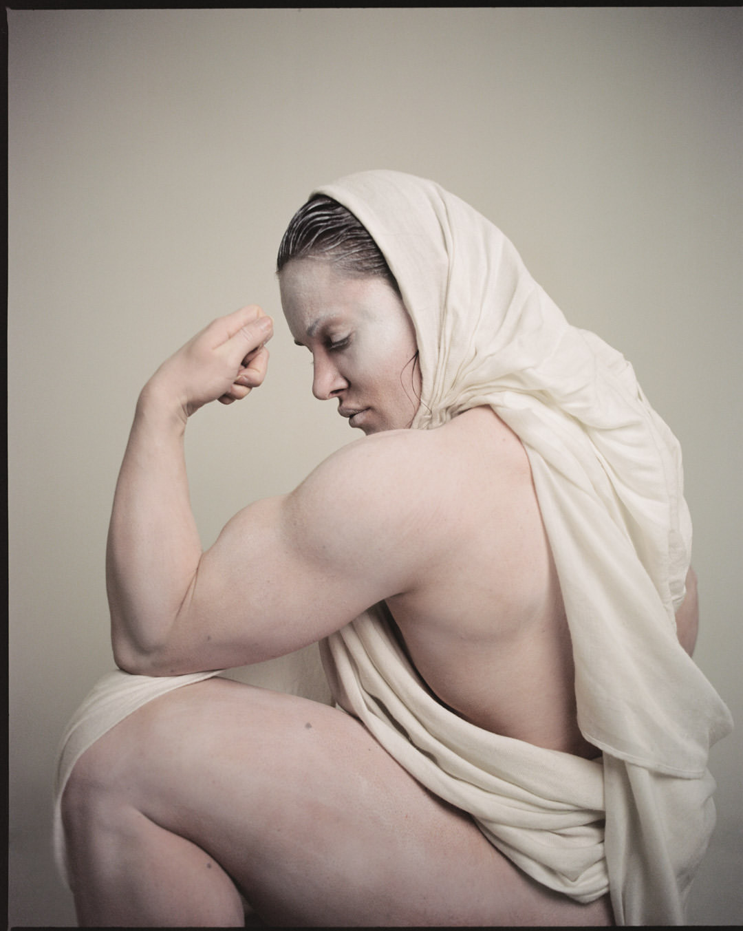 Woman with partially shaved head and body paint draped in white cloth flexing bicep against a neutral background in a studio shot.