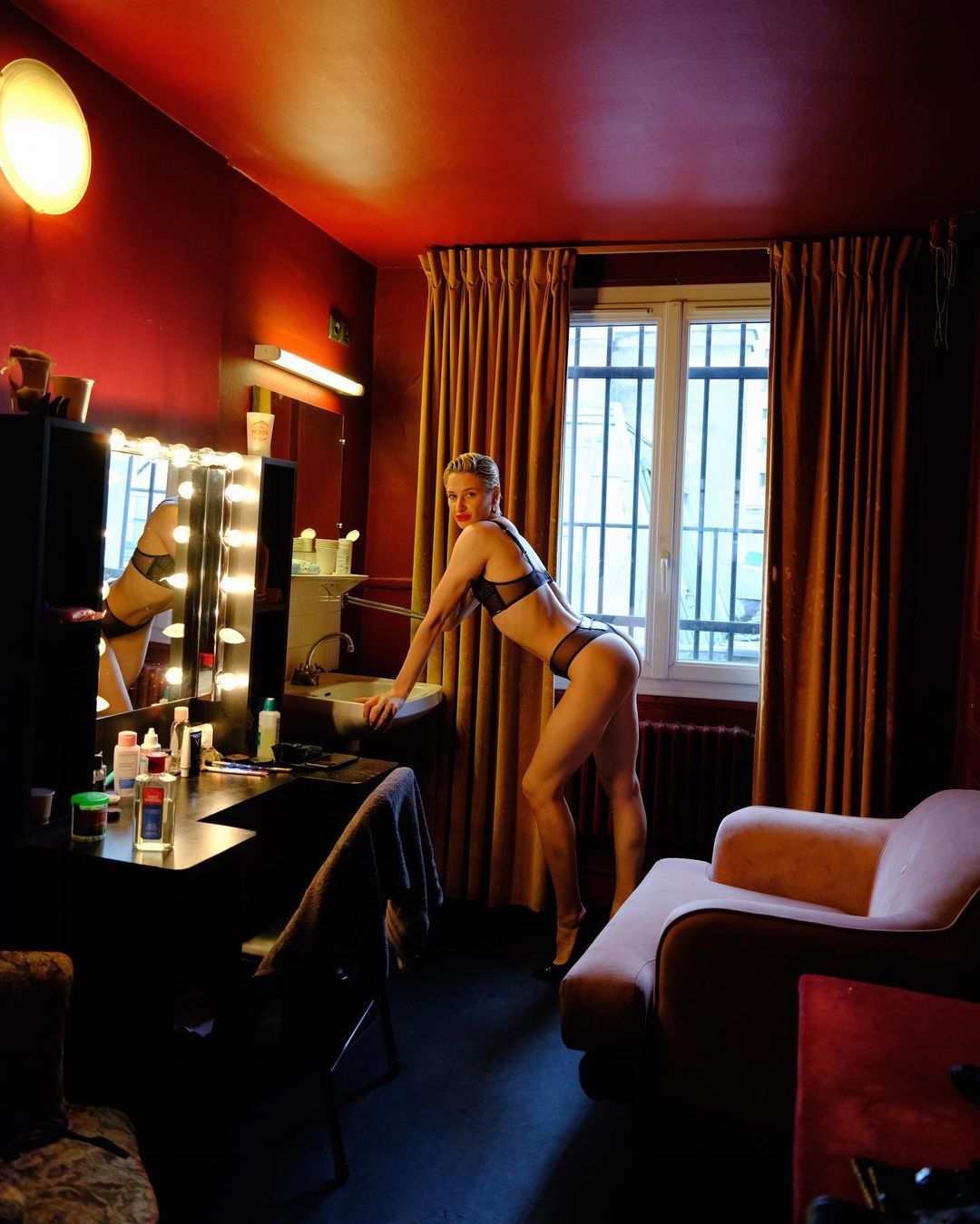 Blonde adult female model in lingerie poses in boudoir setting with vanity mirror, curtains, and a couch inside a dark room.