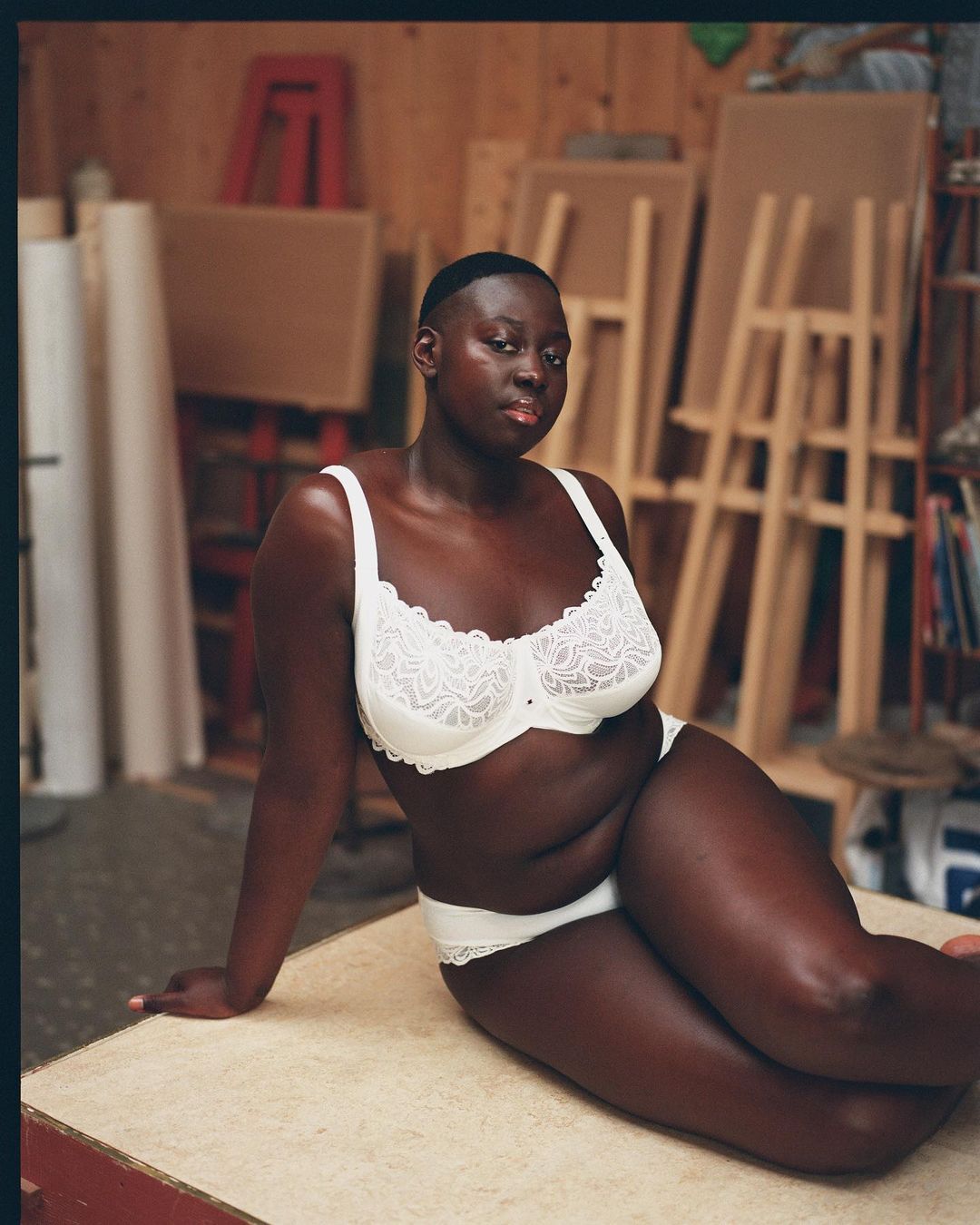 An African-American woman poses indoors in white lingerie, seated on a table against an art studio backdrop featuring easels and art supplies.